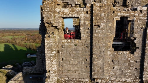 A lady looks out of an unglazed window in an ancient stone wall high up on a ruined castle. In the background can be seen a view across countryside showing that the window is set high in the wall.